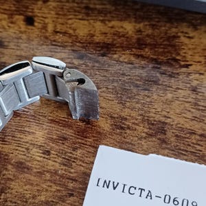 May include: Close-up of a silver-toned watch band clasp. The clasp is made of metal and has a rectangular shape with a textured surface. The text "INVICTA-0609" is printed on a white paper.