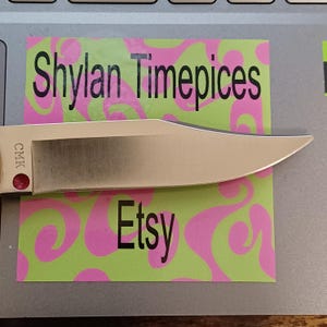 May include: A stainless steel knife blade with a sharp point, resting on a green and pink patterned sticker. The sticker has the text "Shylan Timepices" and "Etsy" in black. The knife handle has a red dot and the letters "CMK".