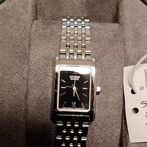 Könnte beinhalten: Eine Citizen Quartz-Uhr mit einem rechteckigen schwarzen Zifferblatt und einem silberfarbenen Edelstahlarmband. Das Zifferblatt zeigt den Markennamen und die römische Ziffer VI. Die Uhr wird in einer grauen Box präsentiert.