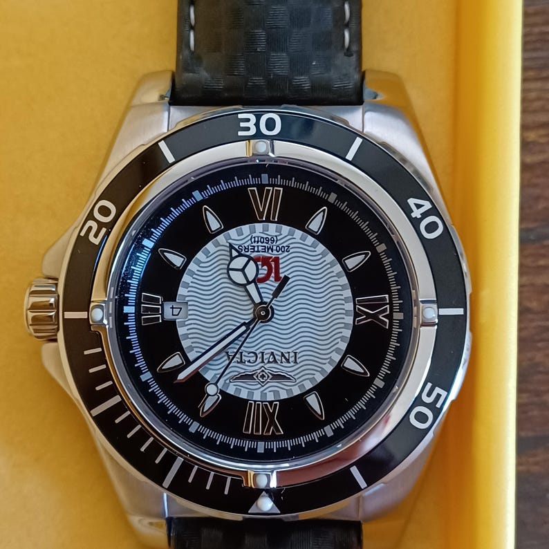 May include: A stainless steel Invicta watch with a black and silver face. The watch features a black bezel with white numerals and a black leather band. The watch face displays the Invicta logo and is water resistant to 200 meters.