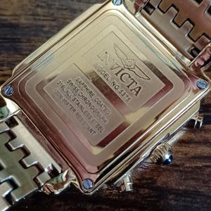 May include: Close-up of a gold-toned Invicta watch back. The watch features the brand name, model number 5375, and details about the sapphire coating, Swiss chronograph, 316L stainless steel, and 100M water resistance.