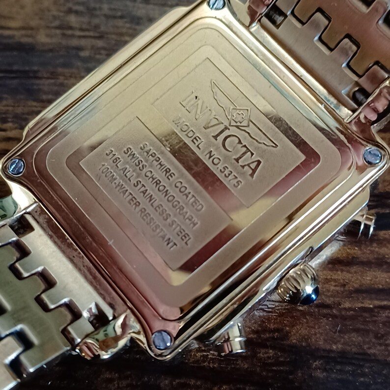 May include: Close-up of a gold-toned Invicta watch back. The watch features the brand name, model number 5375, and details about the sapphire coating, Swiss chronograph, 316L stainless steel, and 100M water resistance.
