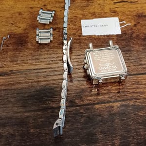 May include: Disassembled Invicta watch components on a wooden surface. The parts include a silver watch band, watch face, and additional links. The watch face has the Invicta logo and text, including "SWISS PARTS CHRONOGRAPH" and "MODEL NO. 0609".