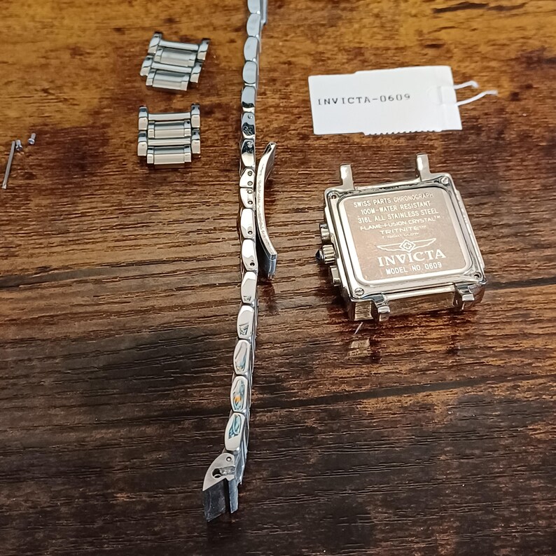 May include: Disassembled Invicta watch components on a wooden surface. The parts include a silver watch band, watch face, and additional links. The watch face has the Invicta logo and text, including "SWISS PARTS CHRONOGRAPH" and "MODEL NO. 0609".