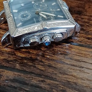 May include: Silver-toned rectangular wristwatch with a blue-grey face. The watch features multiple subdials, a date display, and a bezel encrusted with small, clear stones. The brand name "Invicta" is visible on the face. The watch is set against a dark wooden surface.