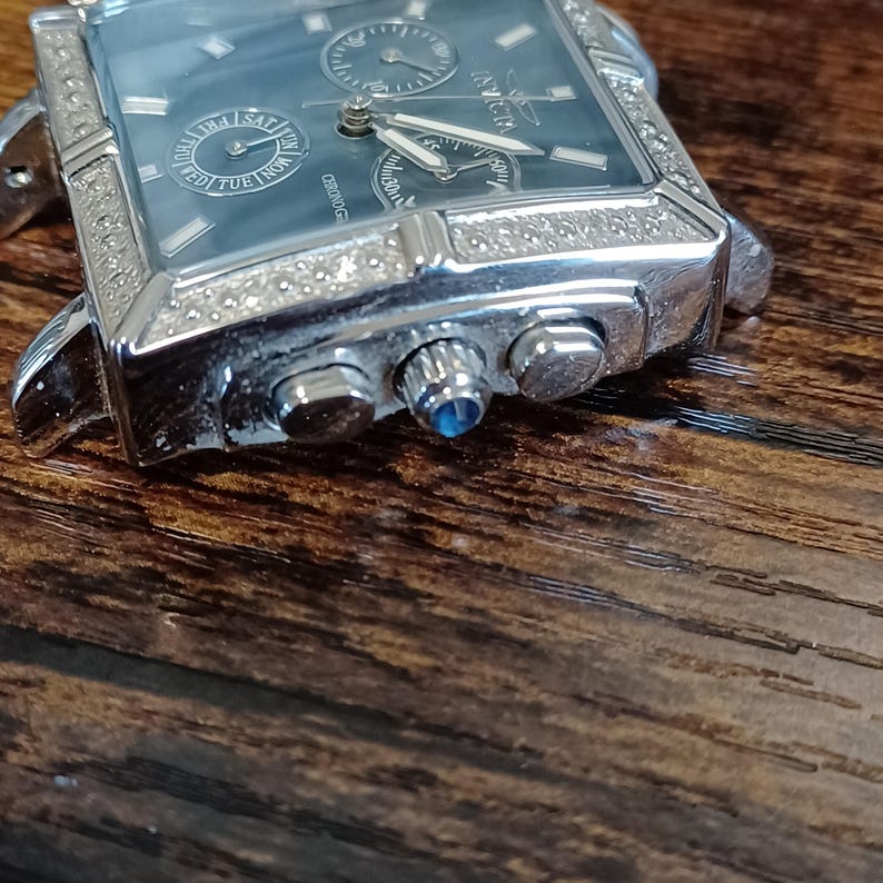 May include: Silver-toned rectangular wristwatch with a blue-grey face. The watch features multiple subdials, a date display, and a bezel encrusted with small, clear stones. The brand name "Invicta" is visible on the face. The watch is set against a dark wooden surface.