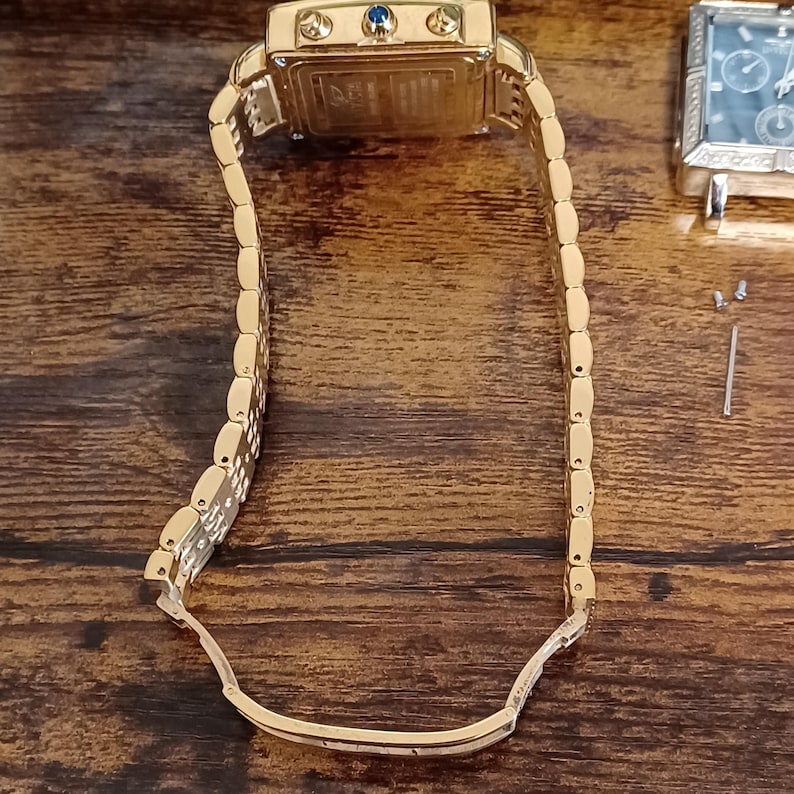 May include: Gold-toned rectangular wristwatch with a matching link bracelet. The watch face has a small blue gem and three smaller dials. The watch is displayed on a wooden surface.