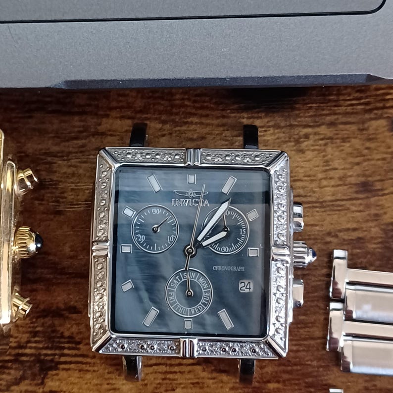May include: Square-faced Invicta chronograph watch with a dark blue dial and silver hands. The watch has a silver bezel with small, clear stones and a black strap. The watch face displays the day, date, and chronograph functions.