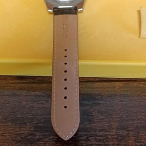 May include: Close-up of a watch with a brown leather strap. The strap is light brown with the words "GENUINE LEATHER" printed on it. The watch face is silver and the background is yellow.
