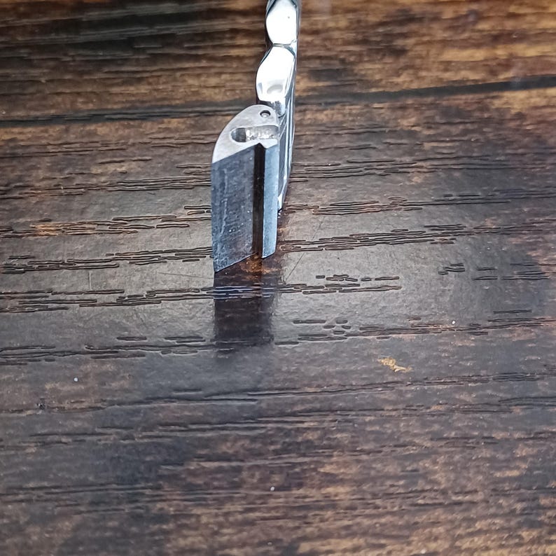 May include: A close-up of a silver metal object with a rectangular shape and a beveled edge. The object is positioned vertically against a dark brown wooden surface, with the light reflecting off the metal.