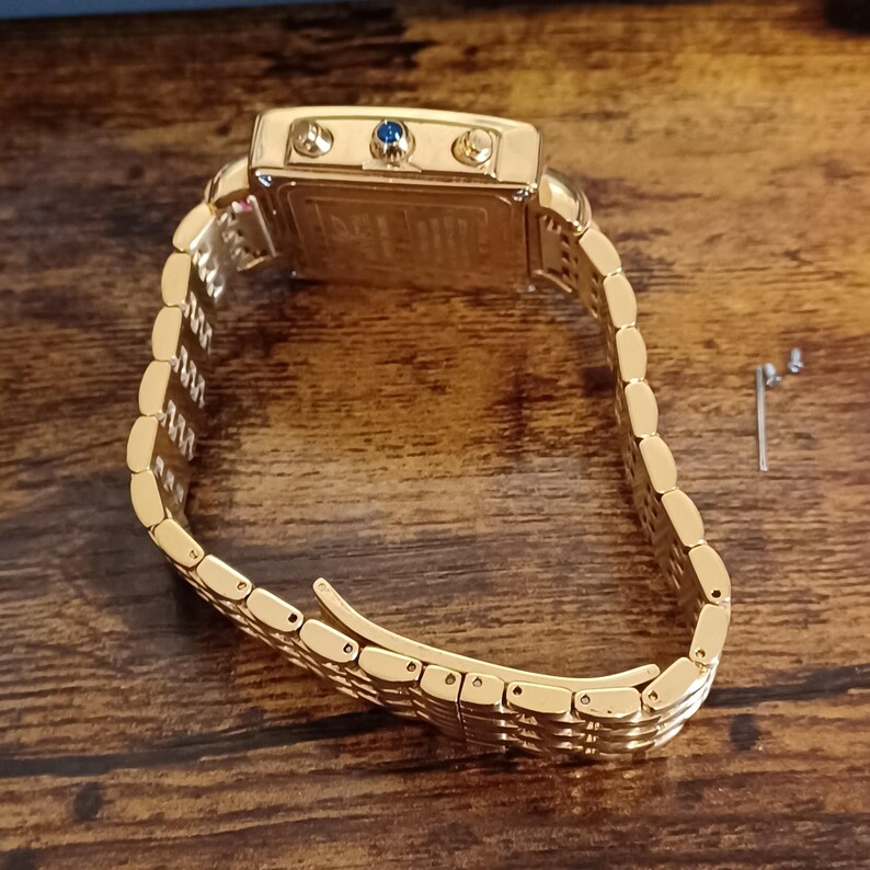 May include: A gold-toned wristwatch with a rectangular face and a matching link bracelet. The watch face features three small dials and a blue gem. The watch is displayed on a wooden surface.