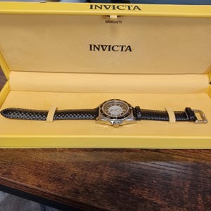 May include: A silver-toned Invicta watch with a black leather band and a patterned face, presented in a yellow box. The watch features a round dial with visible hands and markers. The box is labeled with the Invicta brand name.