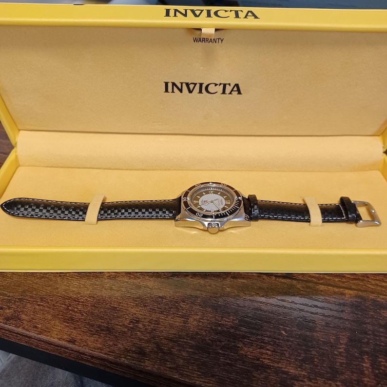 May include: A silver-toned Invicta watch with a black leather band and a patterned face, presented in a yellow box. The watch features a round dial with visible hands and markers. The box is labeled with the Invicta brand name.