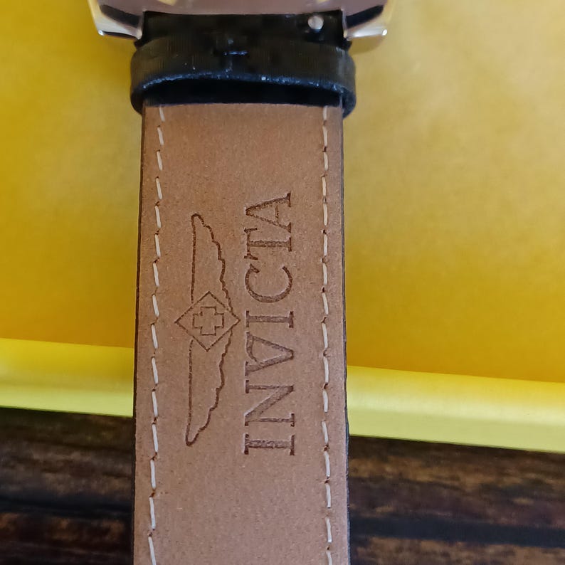 May include: Close-up of a watch strap with the brand name "INVICTA" embossed in brown on a light brown leather strap. The strap is stitched with white thread and has a black leather backing.