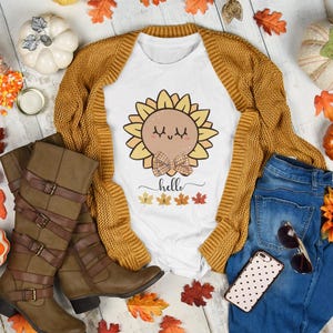 Può includere: Un'immagine a terra a tema autunnale con una maglietta bianca con una grafica di girasole e la parola "hello". Include un cardigan color senape, jeans blu, stivali marroni, zucche e foglie autunnali. Sono presenti anche occhiali da sole e un telefono.