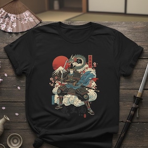 May include: Black t-shirt featuring a Japanese-inspired graphic of a samurai warrior with a dragon, a red sun, and Mount Fuji. The design includes cherry blossoms and Japanese text. The shirt is displayed on a wooden surface with a decorative fan and a katana.