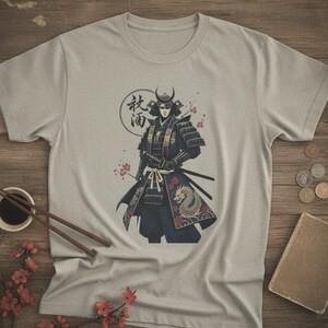 Può includere: T-shirt grigio chiaro con un'illustrazione dettagliata di un guerriero samurai con armatura tradizionale. Il design include un drago, fiori di ciliegio e testo giapponese all'interno di un cerchio. Il samurai porta una spada.