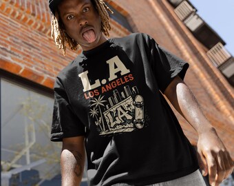 May include: Black t-shirt with "L.A. LOS ANGELES" text and graphic of palm trees, a cityscape, and a skateboard. The person is wearing a black cap and light-wash jeans. The background includes a brick building and a blue sky.
