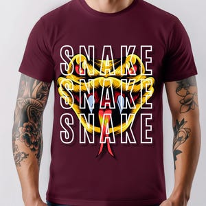 Könnte beinhalten: Ein weinrotes T-Shirt mit einem stilisierten Schlangenmuster. Der Schlangenkopf ist gelb und schwarz mit roten und blauen Akzenten, und das Wort "SNAKE" ist dreimal vertikal in Weiß gedruckt.