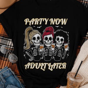 Può includere: T-shirt nero con grafica di scheletri. Il design presenta tre figure di scheletri che tengono in mano delle bevande, con la scritta "PARTY NOW ADULT LATER" in lettere dorate. Gli scheletri hanno acconciature diverse.