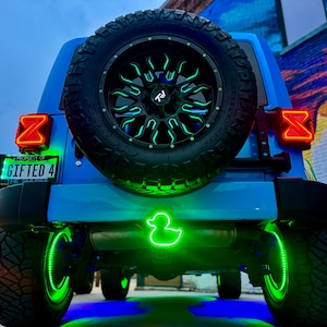 Puede incluir: Vista trasera de un Jeep azul con iluminación de neón verde personalizada. El neumático de repuesto tiene una llanta negra con detalles verdes. La matrícula dice "GIFTED 4". Se ve un pato de neón verde.