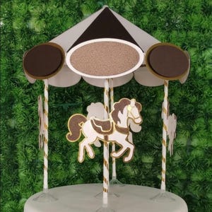 May include: A carousel cake topper with a white and brown canopy, featuring a horse with gold glitter accents. The topper has three oval-shaped signs and is set against a green background.