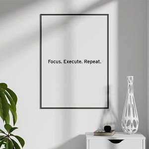 May include: A framed print with the text "Focus. Execute. Repeat." hangs on a white wall. A white decorative vase and a small stack of books sit on a white nightstand. A green leafy plant is in the lower left corner.