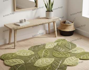 May include: A leaf-shaped rug in shades of green, with detailed vein patterns. The rug is placed on a light-colored wooden floor. A wooden bench, a potted plant, and a woven basket are in the background.