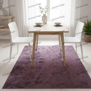 Pode incluir: Um tapete retangular cor de ameixa está centrado sob uma mesa de jantar de madeira clara com duas cadeiras brancas. O quarto tem uma sensação brilhante e arejada com paredes e cortinas brancas. Um vaso de flores brancas e uma planta em vaso completam a decoração.