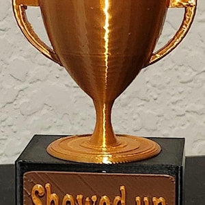 May include: A gold-colored trophy with two handles, sitting on a black base. The base has a brown plaque with the words "Showed up" in gold lettering. The trophy is a symbol of achievement.