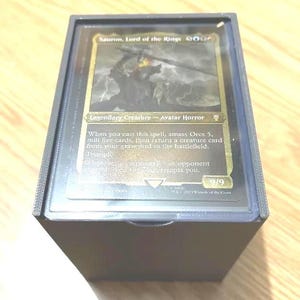 May include: A black card holder containing a Magic: The Gathering card. The card features Sauron, Lord of the Rings, with a dark illustration and text. The card is protected within a clear plastic sleeve.