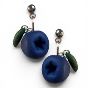 Può includere: Un paio di orecchini pendenti con ciondoli a forma di mirtillo blu opaco, caratterizzati da un'indentazione a forma di stella sulla parte superiore. Ogni mirtillo è decorato con una piccola foglia verde curva fissata a un perno in metallo argentato.