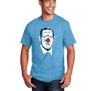 Puede incluir: Camiseta azul claro con un retrato de estilo pop art de una persona con una nariz de payaso roja. El gráfico está en tonos de azul, negro y blanco. La camiseta es de cuello redondo.