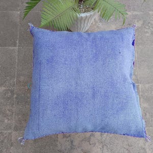 May include: A square, light blue cushion with a textured fabric. The cushion has a stitched border in a darker blue and purple. The corners are finished with small tassels. The cushion is on a gray tiled surface.