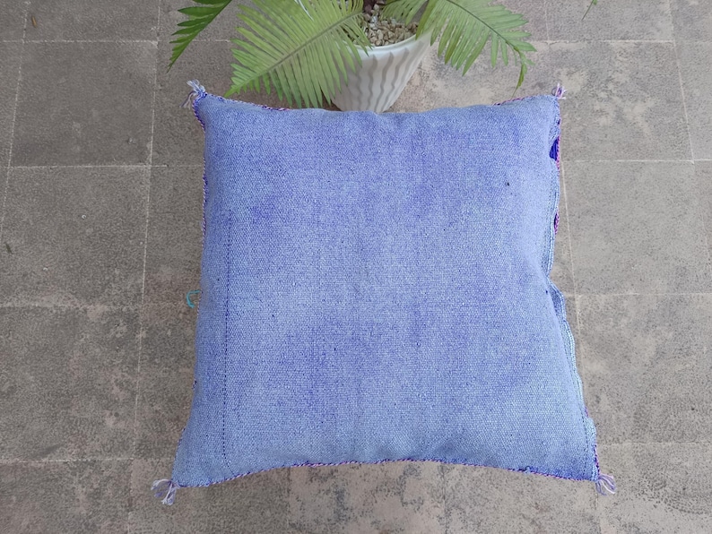 May include: A square, light blue cushion with a textured fabric. The cushion has a stitched border in a darker blue and purple. The corners are finished with small tassels. The cushion is on a gray tiled surface.