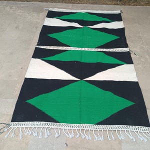 May include: A rectangular woven rug featuring a geometric pattern of large green diamonds, black sections, and off-white triangles. The rug is finished with a white knotted fringe along the top and bottom edges, creating a distinct linear border.