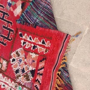 May include: A vibrant, hand-woven rug with a predominantly red background. The rug features intricate geometric patterns in black, white, pink, and blue. The edges are finished with tassels, and the rug is partially folded, revealing a striped section.