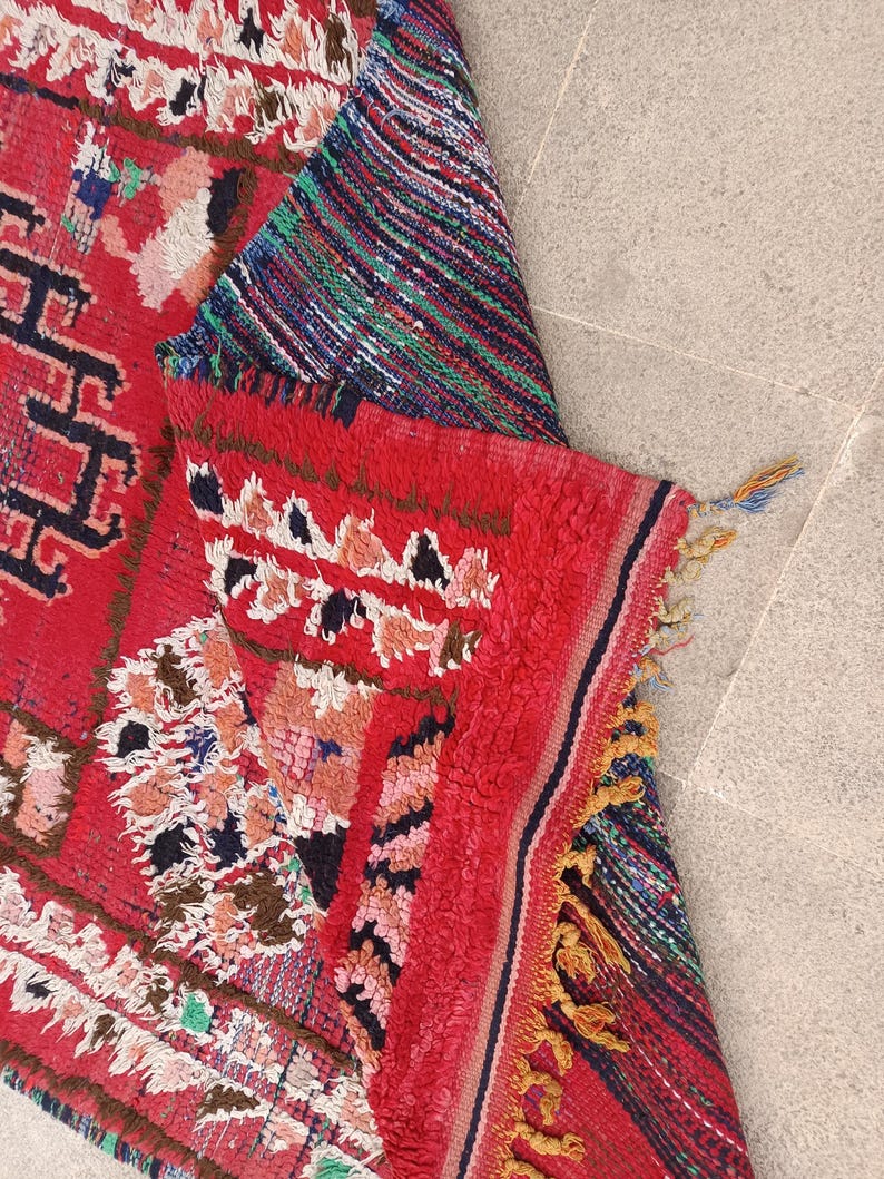 May include: A vibrant, hand-woven rug with a predominantly red background. The rug features intricate geometric patterns in black, white, pink, and blue. The edges are finished with tassels, and the rug is partially folded, revealing a striped section.