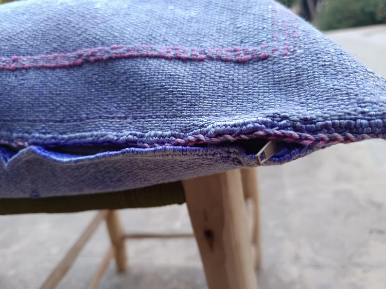 May include: Close-up of a blue woven pillow with a pink stitched design. The pillow has a zipper closure and is resting on a wooden stool. The fabric has a textured appearance.