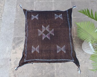 May include: A square, brown woven pillow with a geometric pattern in white and purple. The pillow has a textured surface and is trimmed with a white and blue stitch. The corners have decorative tassels.