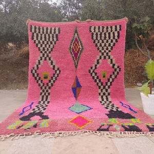 May include: A pink Moroccan rug with geometric patterns. The rug features black and white checkered designs, diamond shapes, and colorful accents. The rug has a fringed edge and is displayed outdoors.