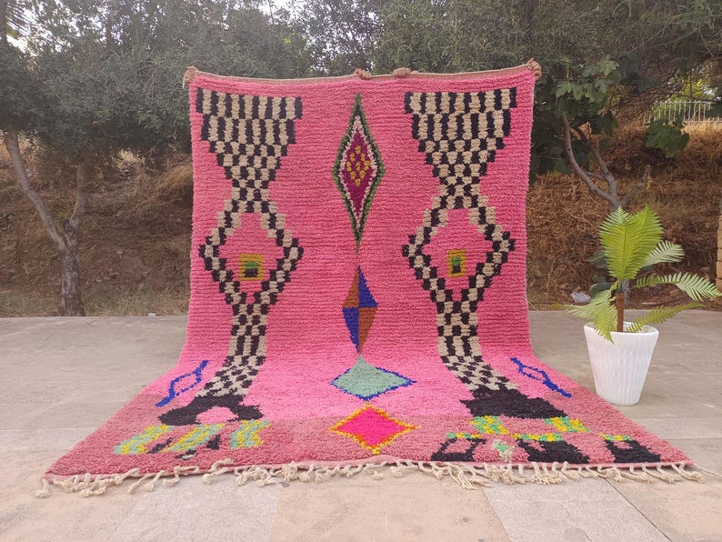 May include: A pink Moroccan rug with geometric patterns. The rug features black and white checkered designs, diamond shapes, and colorful accents. The rug has a fringed edge and is displayed outdoors.