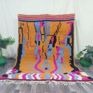 May include: A vibrant, hand-woven rug with a geometric design. The rug features a warm orange background with black, blue, pink, and white abstract shapes. The rug has a fringed edge and is suitable for a living room or bedroom.