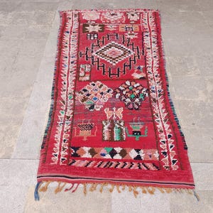 May include: A rectangular red rug with intricate geometric patterns in white, black, and various colors. The rug features a central diamond design and decorative borders, with fringed edges. This handmade rug is suitable for home decor.
