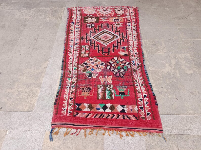 May include: A rectangular red rug with intricate geometric patterns in white, black, and various colors. The rug features a central diamond design and decorative borders, with fringed edges. This handmade rug is suitable for home decor.
