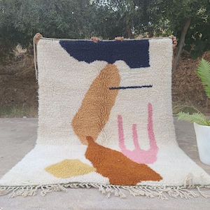 May include: Cream-colored, textured rug with abstract shapes in navy, brown, orange, pink, and yellow. The rug has a fringed edge and is displayed outdoors. The design features a mix of geometric and organic forms, creating a modern aesthetic.