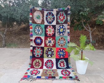 May include: A colorful, hand-woven rug with a patchwork design. The rug features a variety of geometric patterns in squares, with a mix of colors including red, blue, green, and brown. The rug has a fringed edge and is displayed outdoors.