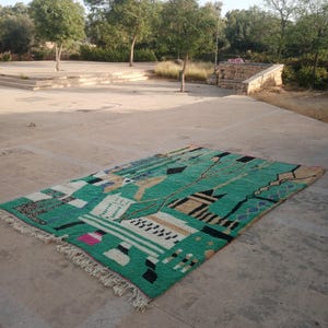 May include: A rectangular, hand-woven rug with a vibrant green background and geometric patterns in black, white, beige, and pink. The rug has a fringed edge and is displayed outdoors on a stone surface.