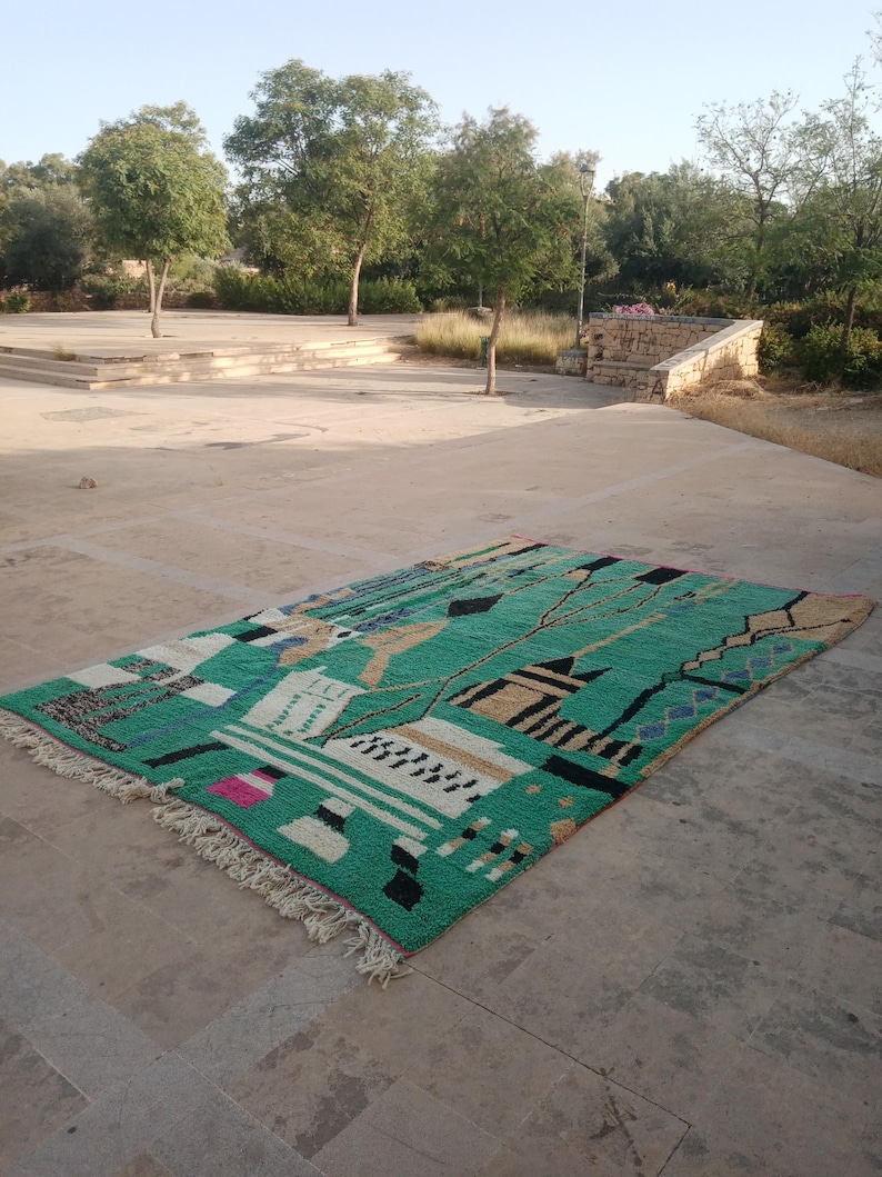 May include: A rectangular, hand-woven rug with a vibrant green background and geometric patterns in black, white, beige, and pink. The rug has a fringed edge and is displayed outdoors on a stone surface.