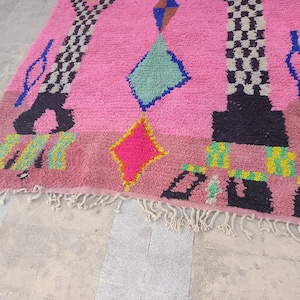 May include: A pink Moroccan rug with geometric designs. The rug features a central pink field with black, blue, yellow, and green geometric shapes. The rug has a fringe along the bottom edge.
